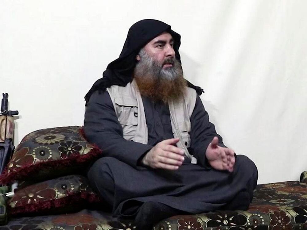  A Muslim man with a long beard and wearing a black turban is sitting on a couch and talking with his hands.