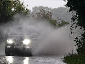 A car with its lights on drivers through a wet road, creating large amounts of spray