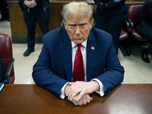 Donald Trump in blue suit and red tie, seated at a wooden table, looks upwards towards the camera