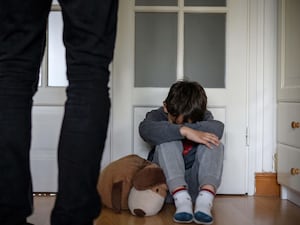 A male child crouched with his head down in his bedroom with a stuffed toy next to him and an adult's legs in the foreground