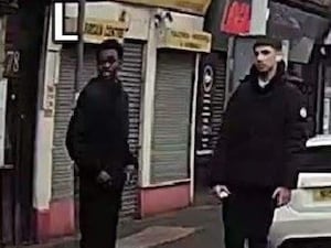 Two young men dressed in black coats standing on a city street in front of shuttered shopfronts