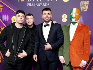 Four men pose in front of the purple bafta backdrop. Two are in black jackets, the one in the middle wears a navy blue tux and bow tie and the fourth wears a suit that is half green and half orange, his face is covered by a balaclava styled after the Irish flag