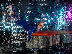 Sir Elton John wearing a blue jumpsuit and doing a handstand on a red baby grand piano