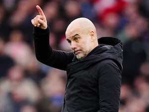 Pep Guardiola, wearing a black coat, points upwards