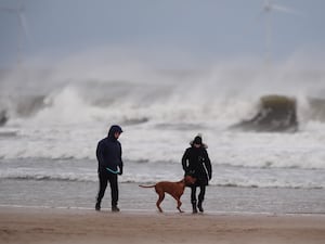 People walking a dog on a beach with large waves and spray in the background