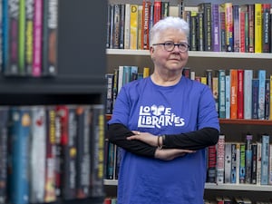 Val McDermid standing with arms folded in front of shelves of books, dressed in a blue t-shirt with Love Libraries written on it in white